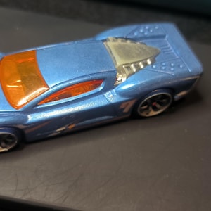 May include: A blue toy race car with orange accents and the number 4 on the front. The car has a silver engine detail and black wheels. The car is on a dark surface.