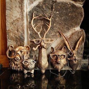 Puede incluir: Colección de bustos de animales en bronce, incluyendo un oso, un gato, un ciervo, un zorro y un conejo. Cada busto lleva gafas. El ciervo tiene una pipa. El conejo tiene un monóculo y un bigote. Los bustos están frente a una pared de piedra.