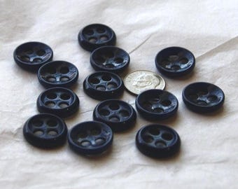 May include: A collection of navy blue buttons with four holes, arranged on a white surface. A U.S. quarter is included for size comparison. The buttons have a slightly domed shape and a glossy finish. Ideal for sewing and craft projects.