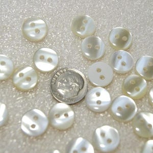 May include: A collection of round, white, two-hole buttons with a pearlescent finish, scattered alongside a silver-coloured United States dime for scale. Each button features a smooth, slightly domed surface and two centred holes for attachment.