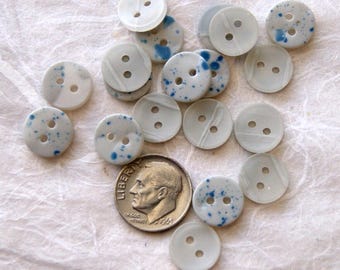 May include: A collection of round, two-hole buttons in white and off-white, some featuring blue speckled patterns or subtle gray lines. A silver-colored United States dime is positioned among the buttons to provide a clear scale reference.