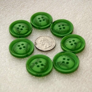 May include: A collection of seven green buttons with four holes each, arranged in a circle. The buttons have a marbled appearance and are next to a U.S. quarter for size comparison. The buttons are on a white surface.