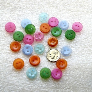 May include: A collection of small, round, translucent plastic buttons in various colors including pink, green, orange, and blue, each featuring two center holes and a radial pattern. A silver United States dime is included for scale.