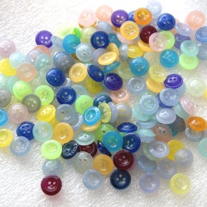 May include: A collection of small, colorful buttons in various shades of blue, yellow, green, purple, and pink. The buttons have two or four holes and a slightly raised edge. Some buttons have a smiling face design.