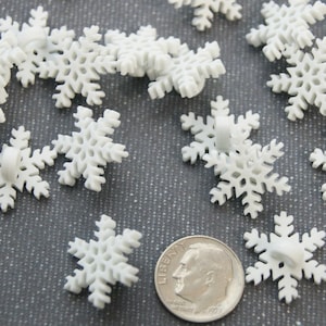 May include: A collection of white snowflake-shaped buttons with intricate branch details. A silver-colored United States dime coin is placed among the buttons to provide a clear sense of scale for the small items.
