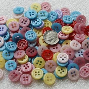 May include: A large assortment of colorful buttons in various shades of pink, blue, yellow, and red. The buttons are made of a smooth, shiny material and have four holes each. There is a US dime coin in the center of the image for scale.