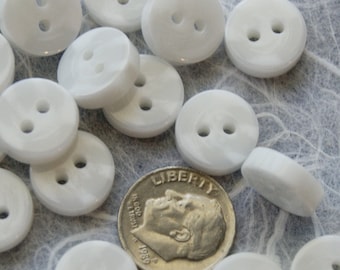 May include: A collection of white plastic buttons with two holes each. The buttons are arranged in a random pattern on a textured surface. A US dime is included for scale.