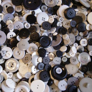 May include: A collection of various sizes and styles of buttons in shades of white, gray, and black. The buttons have two or four holes and are made of plastic. The buttons are scattered, creating a textured surface.