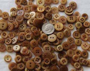 May include: A collection of various sizes of tan and brown buttons with two or four holes. Some buttons have text. A dime is included for size reference. The buttons are scattered on a white surface.