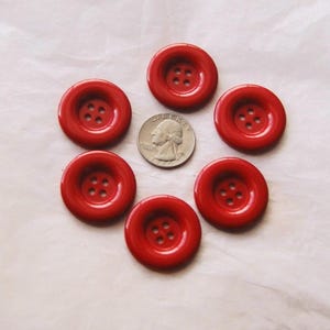 May include: Six bright red buttons with four holes each, arranged in a circle around a quarter. The buttons have a glossy finish and a slightly raised center. These buttons are suitable for sewing projects.