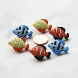 Puede incluir: Seis botones de plástico de colores en forma de pez con contornos negros. Los botones están dispuestos en círculo alrededor de un centavo estadounidense.