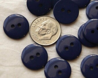 May include: A collection of round, dark navy blue buttons, each featuring two central holes. A silver-colored United States dime is positioned among the buttons to provide a clear visual scale for their size.