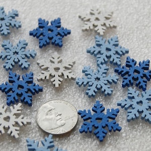 May include: A collection of wooden snowflake ornaments in shades of blue and white. The snowflakes vary in size and design, with some featuring heart-shaped cutouts. A US nickel is included for size reference.