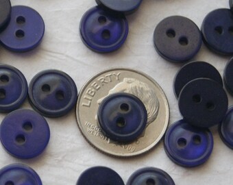 May include: A close-up of a collection of dark blue plastic buttons with two holes each. The buttons are arranged on a white surface with a US quarter for scale.