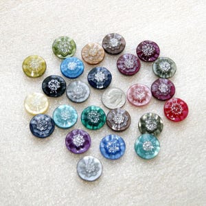 May include: A collection of round, multi-colored buttons featuring a central silver glitter floral pattern. The buttons are arranged in a scattered layout alongside a single United States dime for scale, showcasing various hues including blue, green, red, and purple.