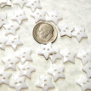 May include: A collection of small, white star-shaped buttons with two holes for sewing. A U.S. dime is included for size comparison. The buttons are scattered across a light-colored surface.