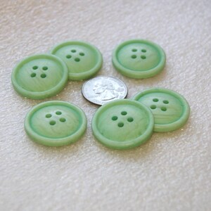 May include: Six light green buttons with four holes each are arranged around a US quarter. The buttons have a slightly marbled appearance and a rounded edge. The quarter is silver and shows a profile of a person.
