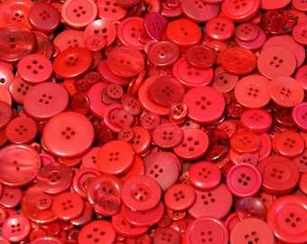 May include: A large collection of assorted red buttons in various sizes and shades, all featuring four holes in the center. The buttons are scattered together in a dense pile, displaying different textures and finishes across the group.
