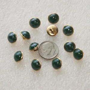 May include: A collection of eleven round, dark green buttons with gold-toned metal rims and shanks. A United States dime is positioned in the center of the buttons to provide a clear scale reference for their size.