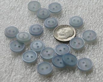 May include: A collection of 17 light blue plastic buttons with two holes each. The buttons are arranged in a random pattern on a white surface. A US dime coin is included for scale.