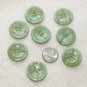 May include: Eight round, sage green buttons with a marbled, translucent finish and four central holes. Each button features a smooth, glossy surface. A standard United States quarter coin is placed among the buttons for scale.
