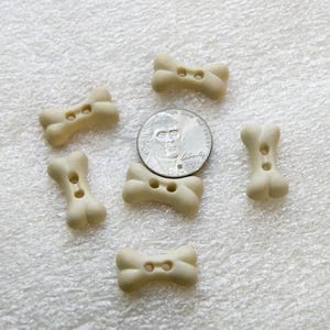 May include: Five off-white bone-shaped buttons, each featuring two central holes for attachment. A silver-colored United States nickel coin is positioned in the center of the arrangement to provide a scale reference for the size of the buttons.