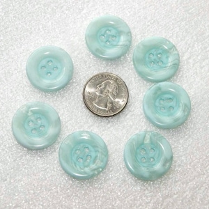 May include: Eight round, light blue buttons with four holes each. The buttons have a marbled appearance with white swirls. A US quarter is included for size comparison. The buttons are arranged in a circle.