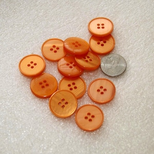 May include: A collection of orange, round buttons with four holes each. The buttons are made of a glossy material and are scattered on a white surface. A US quarter is included for size comparison.