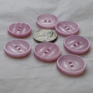 May include: A collection of light pink, oval-shaped buttons with two holes each. The buttons have a striped design with the word "refined" printed on them. A silver coin is placed in the center of the buttons.