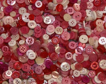 May include: A large collection of assorted buttons in various shades of pink and red. The buttons are made of plastic and have a smooth, shiny finish.