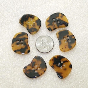 Peut inclure: Cinq boutons irréguliers en forme de rein avec un motif écaille de tortue marron et noir, chacun doté de quatre trous centraux. Une pièce de monnaie américaine de 25 cents est placée au centre pour l'échelle, mesurant 24,26 mm de diamètre.