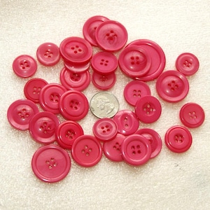 May include: A collection of various sizes of bright pink buttons with four holes. A U.S. quarter is included for size comparison. The buttons are made of a smooth, glossy material and are scattered on a white surface.