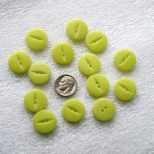 May include: A collection of light green, round buttons with a textured surface and a slit design. Each button has two holes for sewing. A U.S. dime is included for size comparison. The buttons are scattered on a white surface.