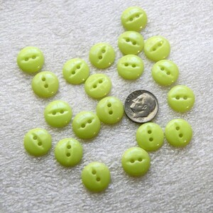 May include: A collection of twenty small, round, lime green buttons, each featuring two central holes. A silver-colored United States dime is positioned among the buttons to provide a sense of scale for the size of the individual items.