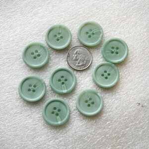 May include: A collection of ten round, light green buttons with four holes each. The buttons have a marbled appearance and are arranged around a quarter coin for size comparison. The buttons are approximately 1 inch in diameter.