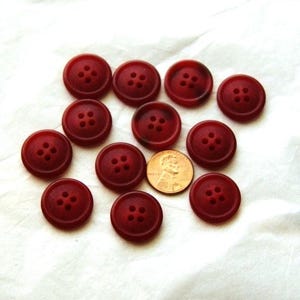May include: A collection of eleven round, dark red buttons with four holes each. A penny is included for size comparison. The buttons are arranged on a white, textured surface. The buttons are a solid color.