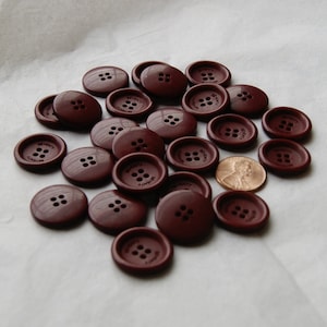 May include: A collection of dark brown buttons with four holes, scattered on a white surface. Each button is round with the word "SEVENTY" printed on it. A penny is included for size comparison.