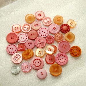 May include: A collection of assorted sewing buttons in various shades of pink, coral, and orange. The buttons feature different shapes including round and square, with varying textures, patterns, and hole counts, scattered across a textured surface.