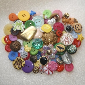 May include: A collection of assorted buttons in various colors, shapes, and sizes. The buttons include round, square, and novelty designs, such as a frog, a pumpkin, and a dice. Colors include red, yellow, green, blue, and purple.
