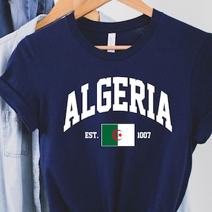 以下が含まれることがあります： 「ALGERIA EST. 1007」という白い文字とアルジェリアの国旗がプリントされたネイビーブルーのTシャツです。