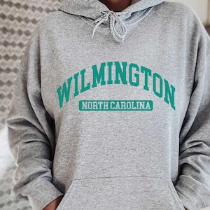 Op de afbeelding: Grijs hoodie met de tekst "WILMINGTON NORTH CAROLINA" in turquoise.