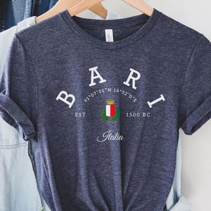 Pode incluir: Uma t-shirt azul marinho com a palavra "BARI" em letras brancas. Abaixo, as coordenadas 41°07'31"N 16°52'0"E, um brasão e a palavra "Italia". A t-shirt está pendurada num cabide de madeira.