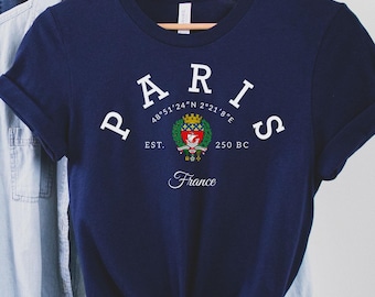 May include: A navy blue t-shirt with the word "PARIS" in white arched letters. Below the word "PARIS" is a crest and the text "EST. 250 BC France". A light blue button-down shirt and a brown hat hang on the rack behind the t-shirt.