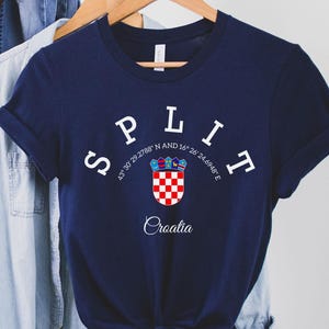 May include: A navy blue t-shirt with the word "SPLIT" arched across the top in white letters. Below the word is a Croatian coat of arms and the word "Croatia". The shirt is hanging on a wooden hanger.