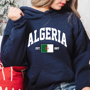 May include: A navy blue hooded sweatshirt with the word "ALGERIA" in white letters across the chest. The sweatshirt also features the Algerian flag and the text "EST. 1007".