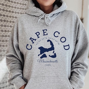 May include: A grey hooded sweatshirt with a front pocket and drawstring. The sweatshirt features the words "CAPE COD" in navy blue arched above a navy blue map of Cape Cod, with the word "Massachusetts" and the year "1685" below the map.