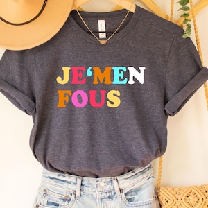 Puede incluir: Una camiseta gris oscuro con el texto "JE'MEN FOU" en letras de colores. La camiseta está colgada de una percha de madera y se combina con pantalones cortos de mezclilla y un bolso de paja.