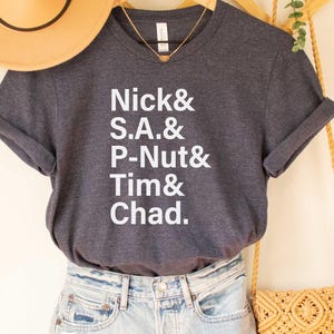May include: A dark gray t-shirt with white text that reads "Nick & S.A. & P-Nut & Tim & Chad."