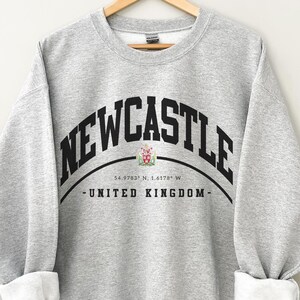 May include: A light gray sweatshirt with the word "NEWCASTLE" arched across the chest in black block letters. Below the text is a crest and the words "UNITED KINGDOM". The sleeves are cuffed, revealing white fabric.