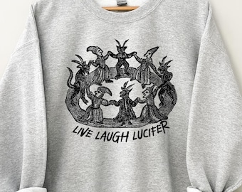 Peut inclure: Un sweat-shirt gris clair avec un motif noir de personnages en cercle, avec l'inscription "LIVE LAUGH LUCIFER" en dessous. Le sweat-shirt a des manches longues avec des poignets retroussés.